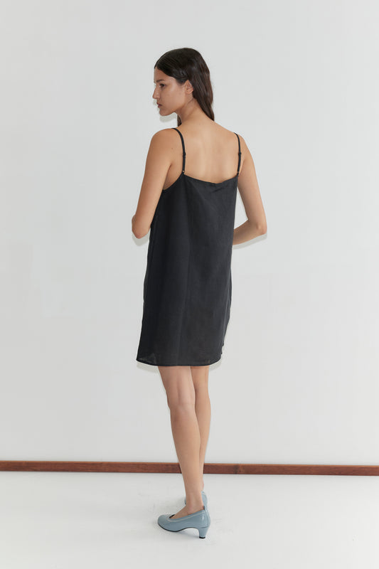 A woman with long dark hair wears the Weave Mini - Black, a simple black linen mini dress with adjustable straps, paired with light blue shoes. She stands indoors against a plain white wall, facing slightly to the side.