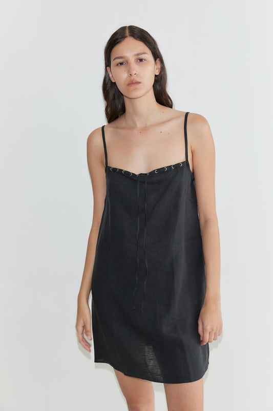 A woman with long dark hair stands against a white background, wearing the Weave Mini - Black dress from Deiji Studios. The black linen mini dress features adjustable straps. She has a neutral expression and her arms rest at her sides.