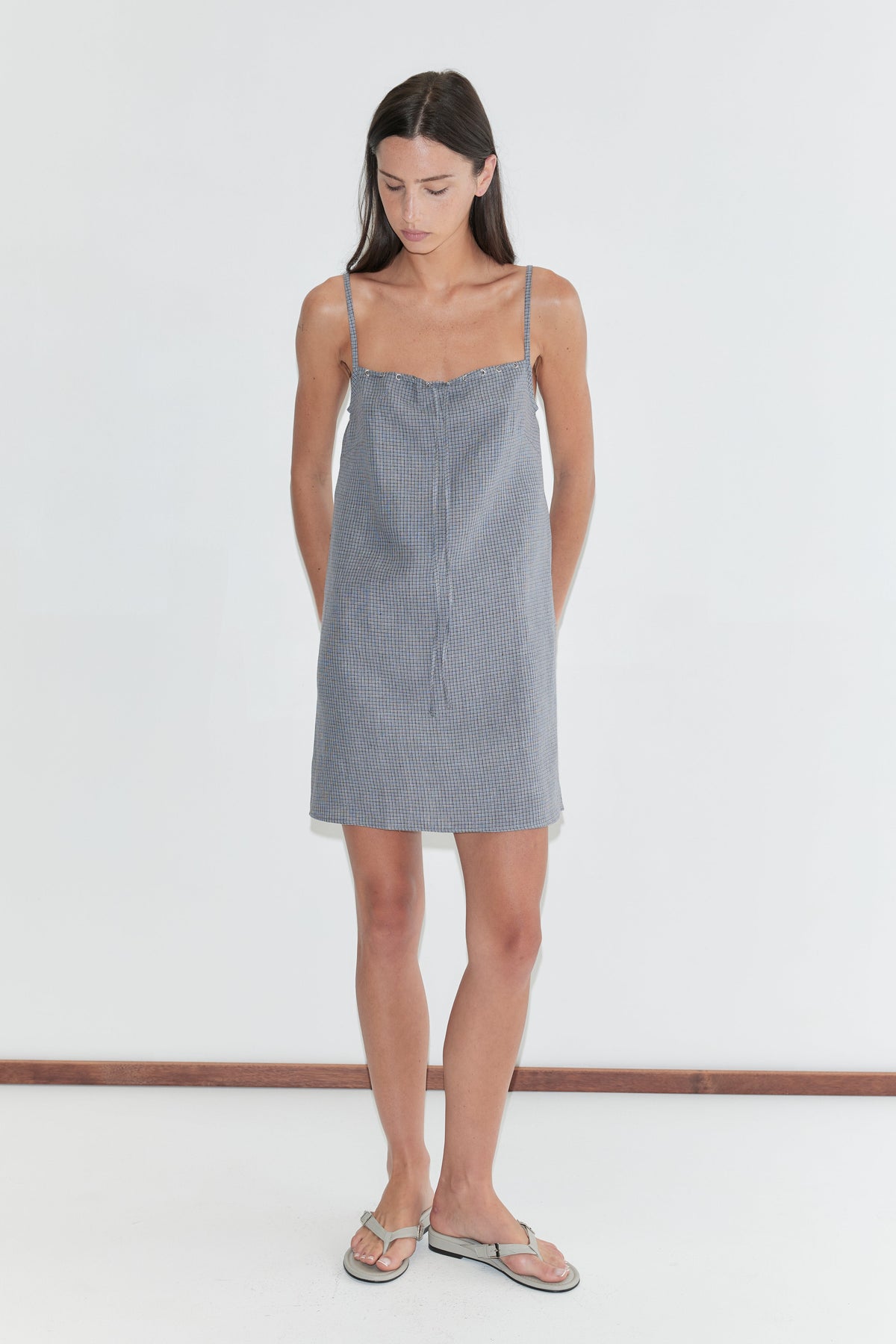 A woman with long dark hair stands against a plain white background looking down, wearing the Weave Mini - Indigo Check by Deiji Studios, an indigo micro-check linen mini dress with thin straps, and flip-flops.