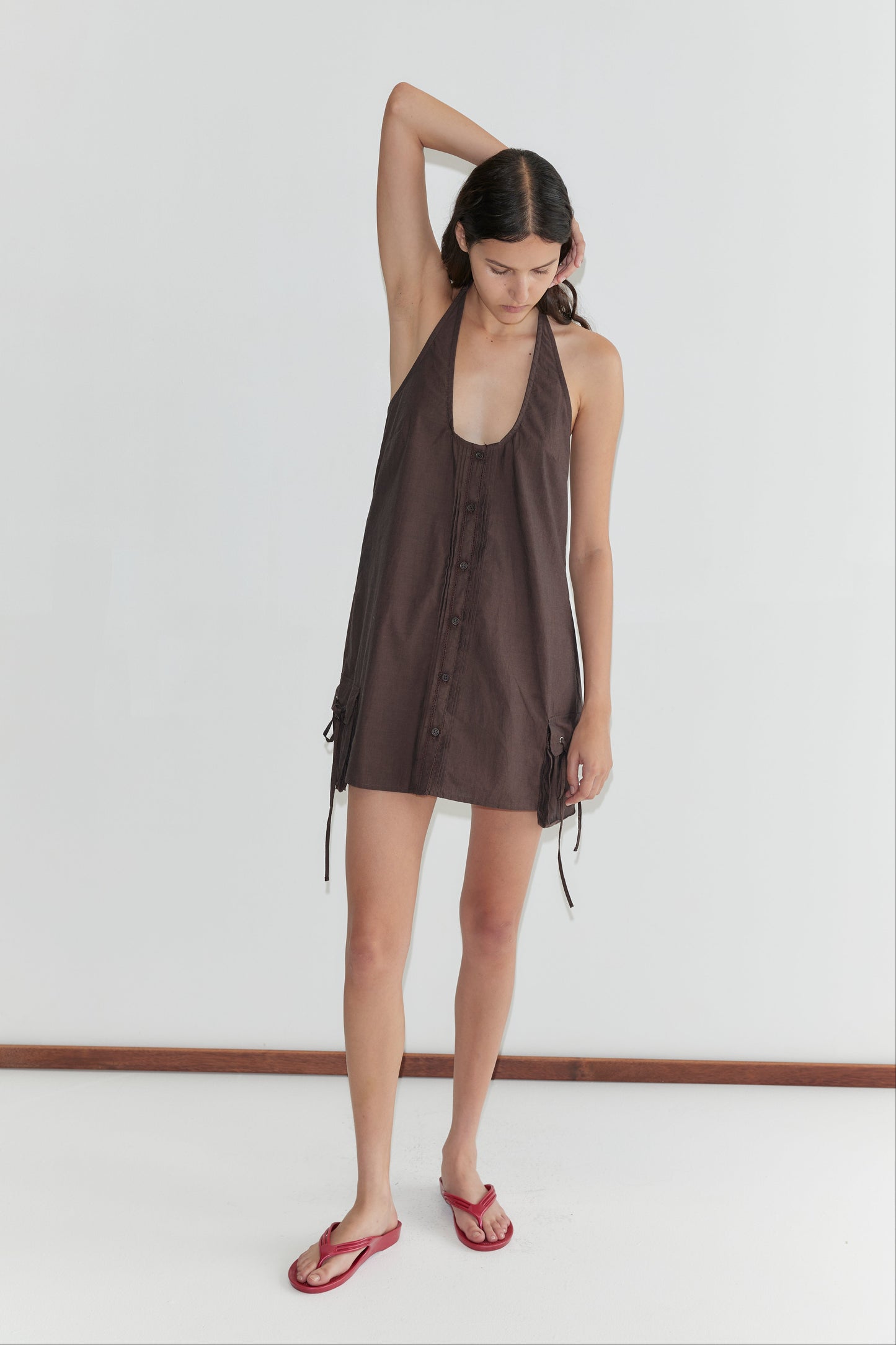 A woman stands against a plain white wall wearing the Pocket Halter Dress - Toast with side ties and red flip-flops. She has one arm bent, hand resting on her head, looking down.