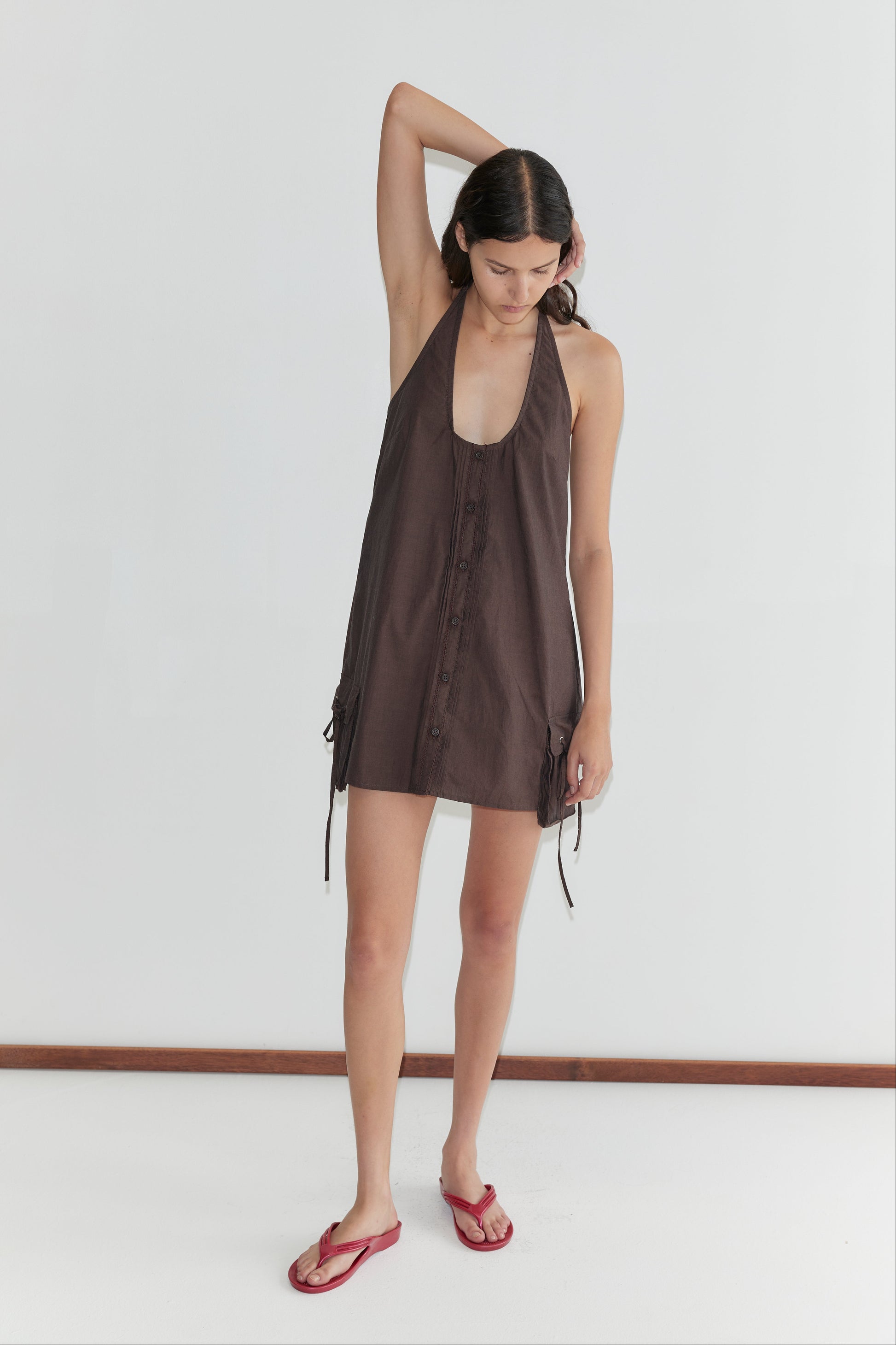 A woman stands against a plain white wall wearing the Pocket Halter Dress - Toast with side ties and red flip-flops. She has one arm bent, hand resting on her head, looking down.