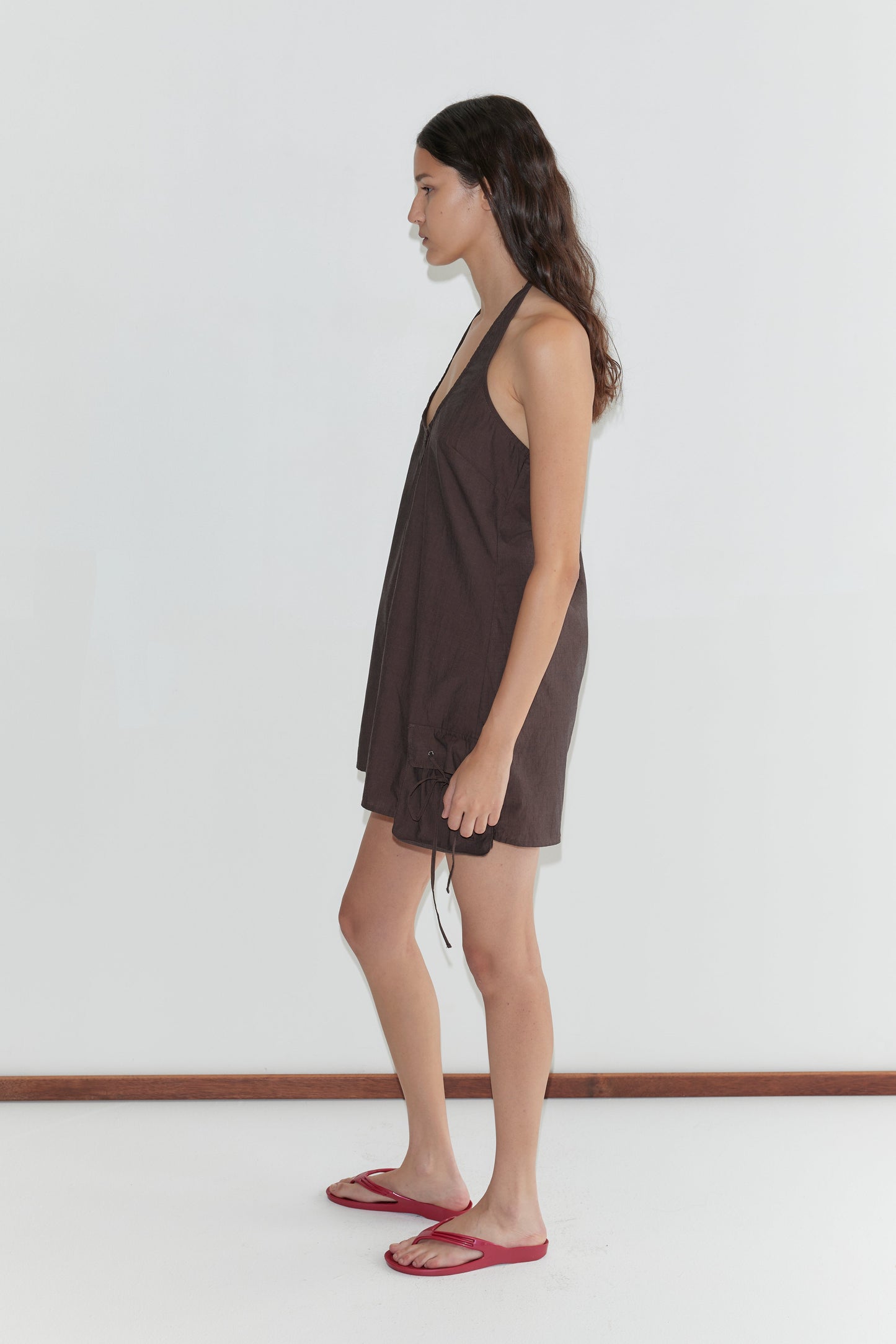 A woman with long dark hair stands sideways against a plain white wall, wearing the Pocket Halter Dress - Toast and red flip-flops.