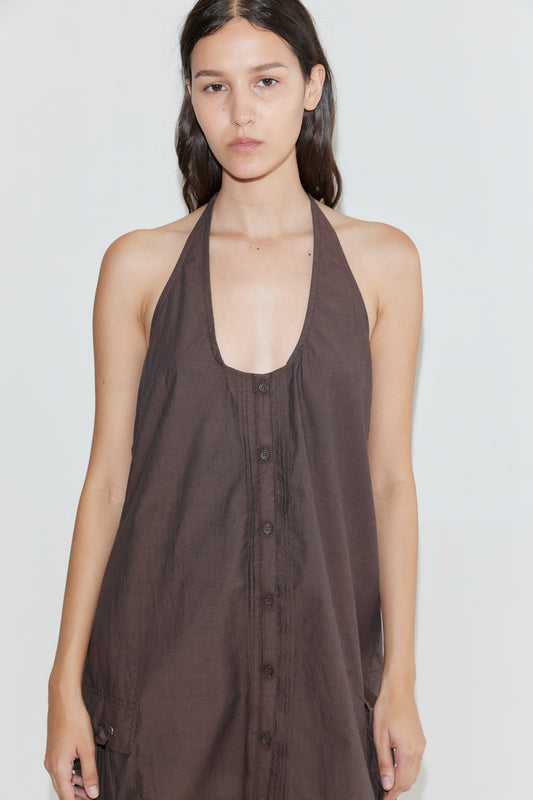 A woman with long dark hair wears the Pocket Halter Dress - Toast, a sleeveless brown adjustable halter dress with front buttons, standing against a plain light background and looking at the camera with a neutral expression.