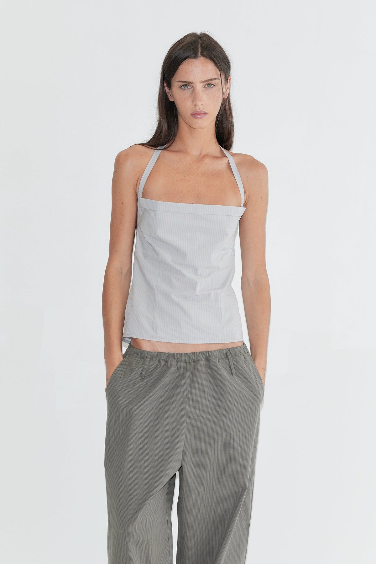 A woman with long dark hair stands against a plain white background wearing the Folded Top - Cloud Stripe by Deiji Studios, a pale grey-blue fine-striped fold-over halter top, with grey check wide-leg trousers and flip-flops.