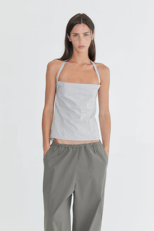 A woman with long dark hair stands against a plain white background wearing the Folded Top - Cloud Stripe by Deiji Studios, a pale grey-blue fine-striped fold-over halter top, with grey check wide-leg trousers and flip-flops.