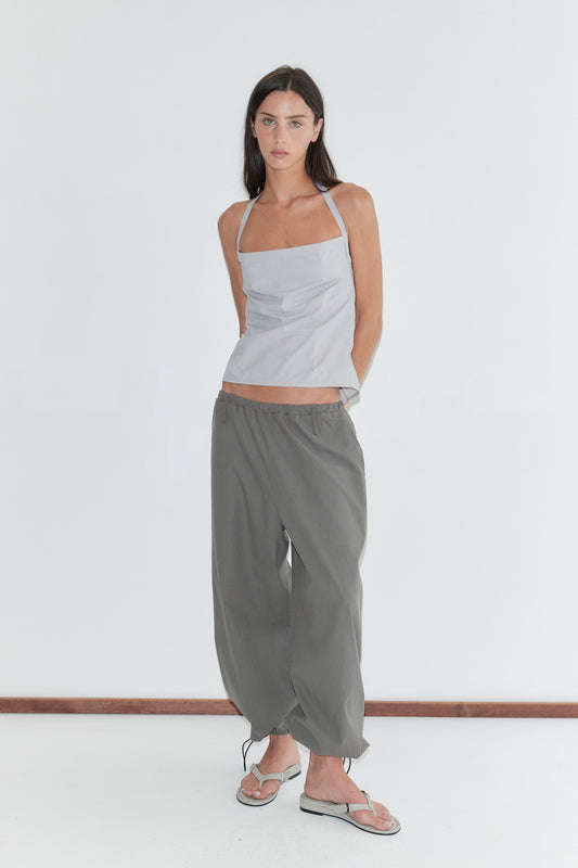 A woman with long dark hair stands against a plain white background wearing the Folded Top - Cloud Stripe by Deiji Studios, a pale grey-blue fine-striped fold-over halter top, with grey check wide-leg trousers and flip-flops.