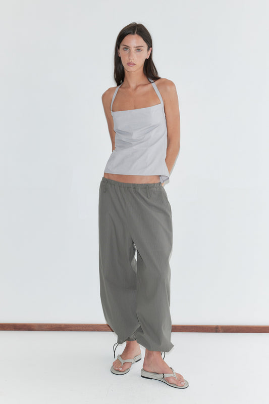 A woman with long dark hair stands against a plain white background wearing the Hem Toggle Pant - Moss Stripe by Deiji Studios, moss-green fine-check trousers with a toggle-gathered hem, with a pale grey striped halter top and flip-flops.