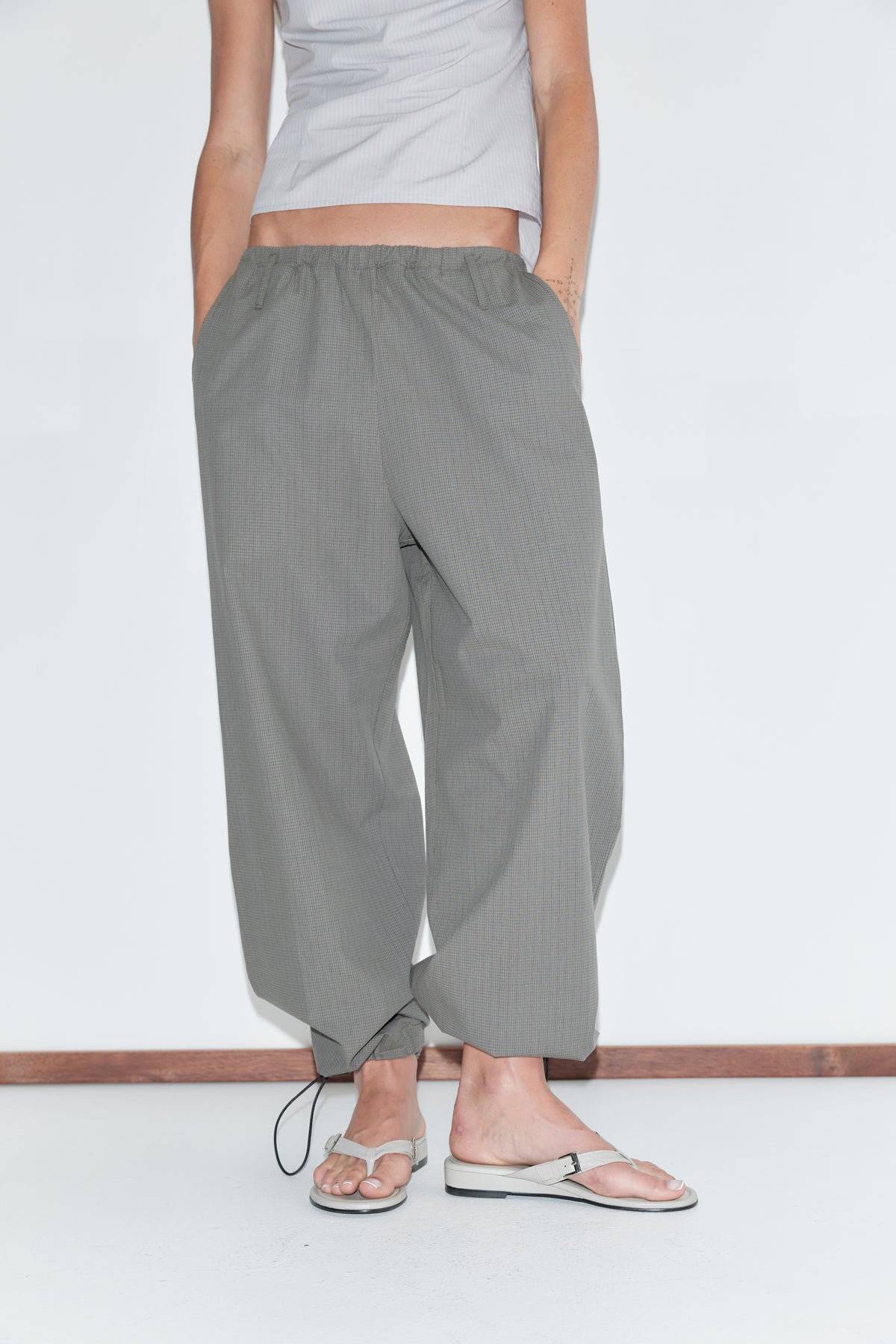 A cropped view of the Hem Toggle Pant - Moss Stripe by Deiji Studios, moss-green fine-check trousers with a toggle-gathered hem, worn with flip-flops against a plain white background.