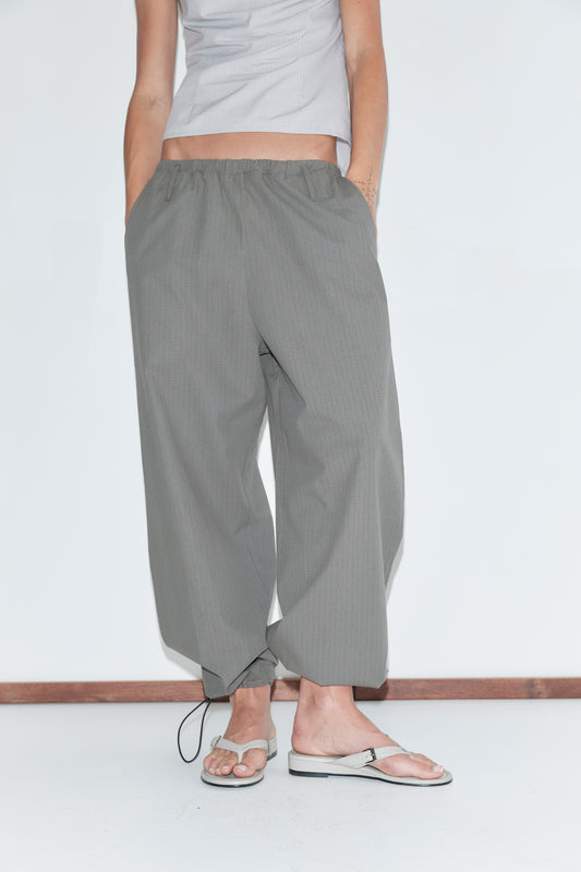 A cropped view of the Hem Toggle Pant - Moss Stripe by Deiji Studios, moss-green fine-check trousers with a toggle-gathered hem, worn with flip-flops against a plain white background.