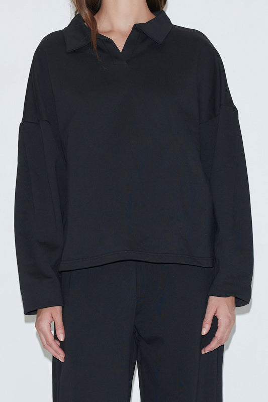 A person models the Jersey Jumper - Black, a loose-fitting cotton piece with an open polo collar, paired with matching black pants. They stand against a plain light background, their head partially out of frame.