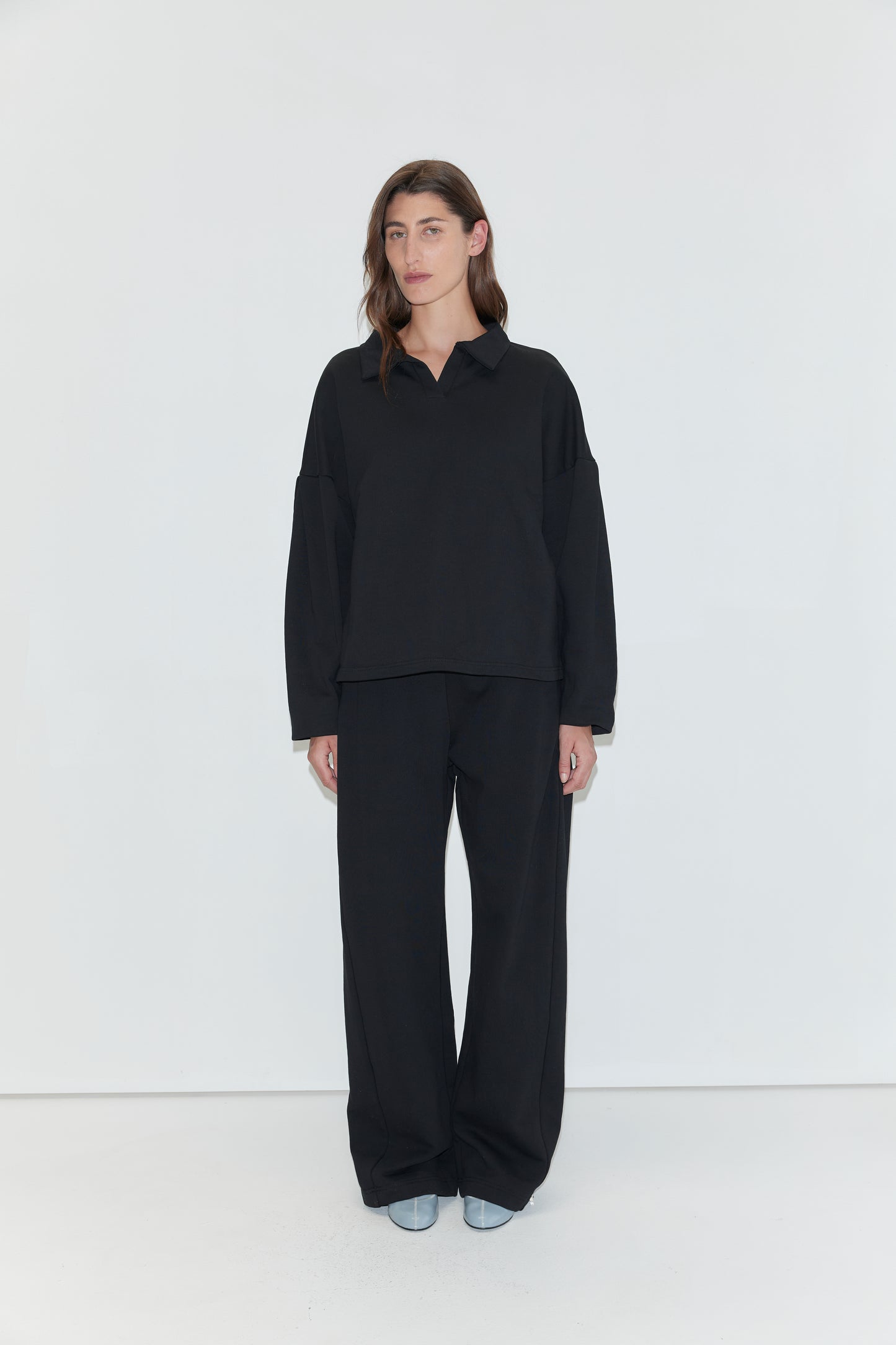 A person with long brown hair is wearing the Jersey Jumper - Black, a loose-fitting cotton piece with an open polo collar, paired with black pants and light-colored shoes, standing against a plain white background.