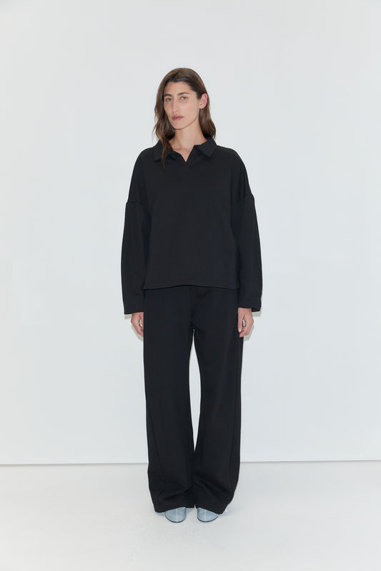 A person with long brown hair is wearing the Jersey Jumper - Black, a loose-fitting cotton piece with an open polo collar, paired with black pants and light-colored shoes, standing against a plain white background.