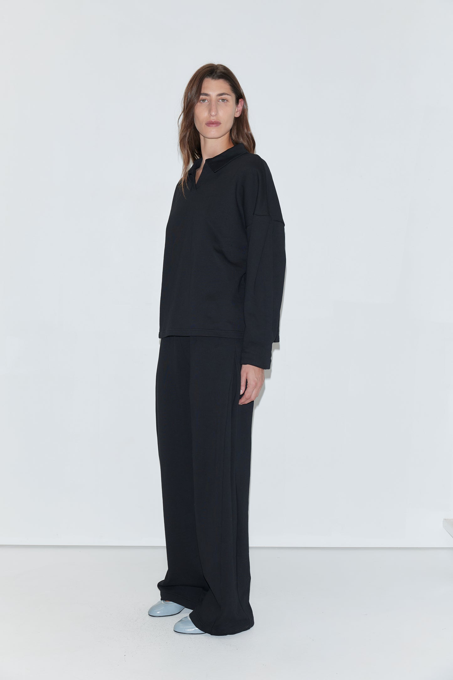 A person with long brown hair stands against a white background, wearing the Jersey Jumper in black—a loose-fitting top featuring an open polo collar—paired with wide black pants and light blue shoes.