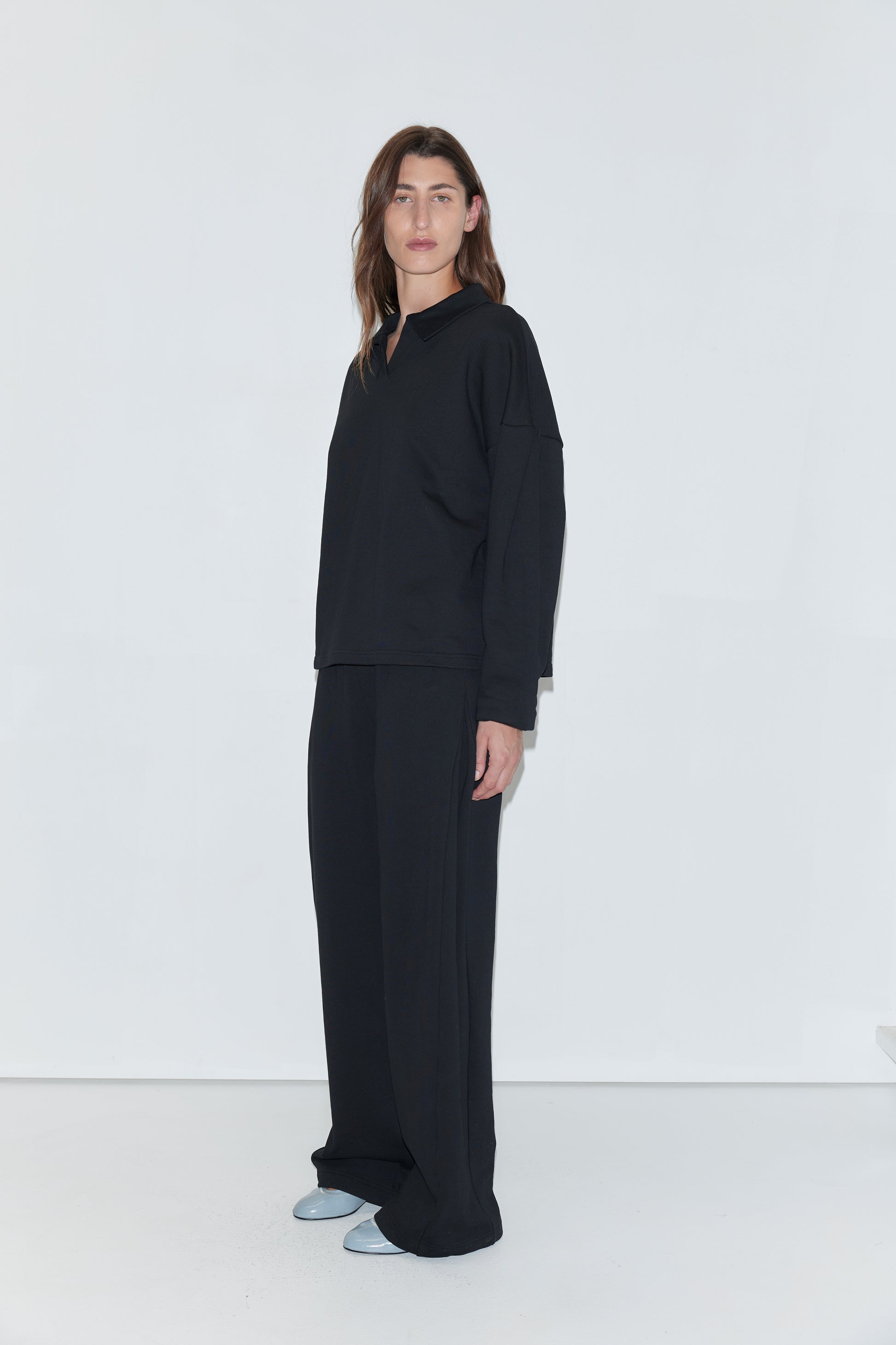 A person with long brown hair stands against a white background, wearing the Jersey Jumper in black—a loose-fitting top featuring an open polo collar—paired with wide black pants and light blue shoes.