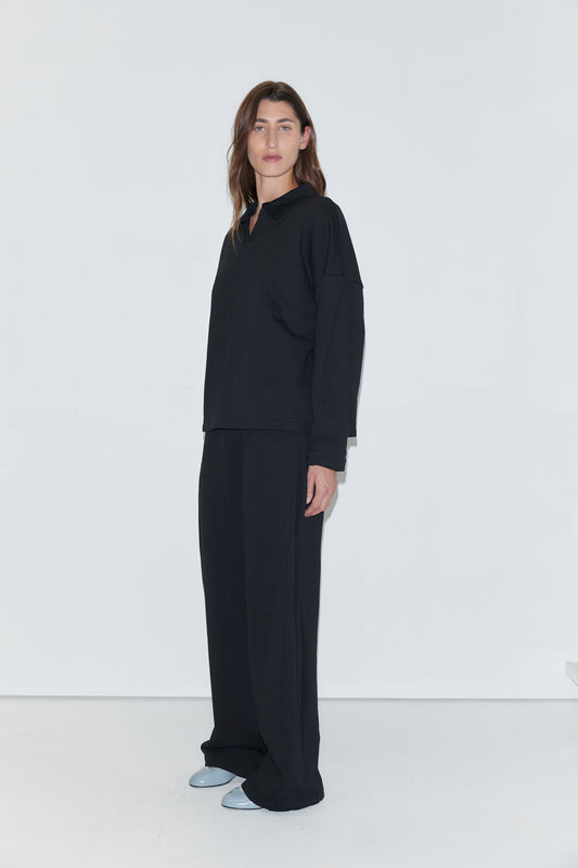A person with long brown hair stands against a white background, wearing the Jersey Jumper in black—a loose-fitting top featuring an open polo collar—paired with wide black pants and light blue shoes.