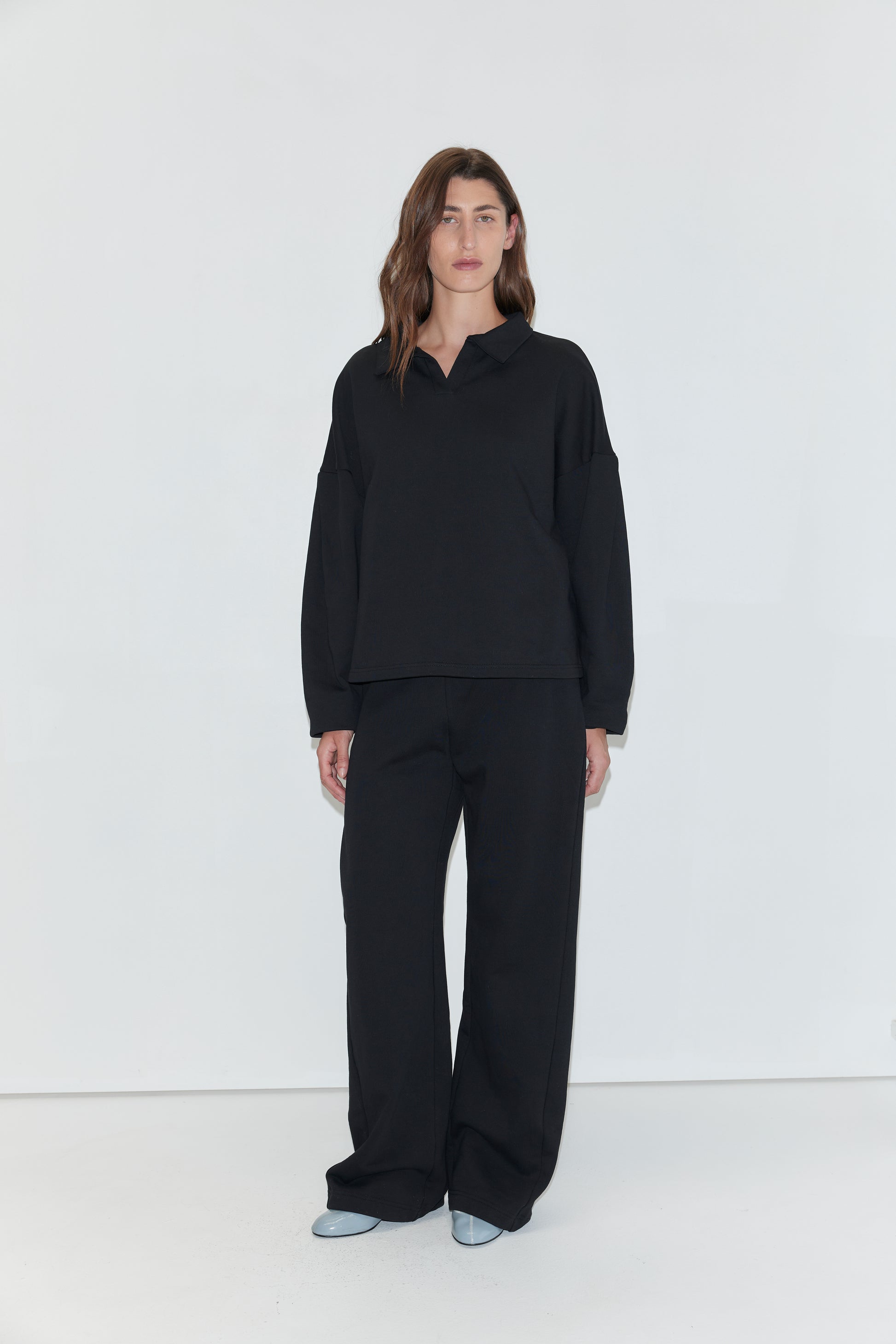 A woman with long brown hair stands facing the camera against a plain white background, wearing a black long-sleeved polo shirt and the Side Pleat Pant - Black from Deiji Studios. She is wearing light blue pointed-toe shoes.