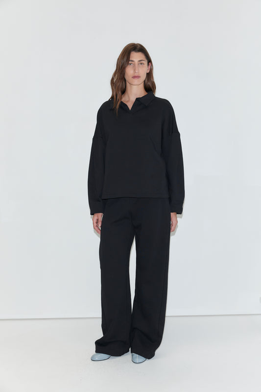 A woman with long brown hair stands facing the camera against a plain white background, wearing a black long-sleeved polo shirt and the Side Pleat Pant - Black from Deiji Studios. She is wearing light blue pointed-toe shoes.