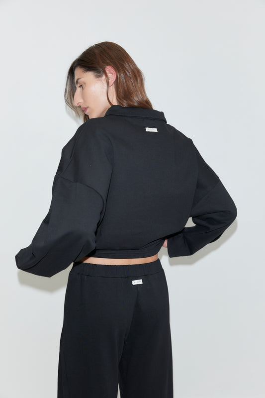 A person with long brown hair is wearing the Jersey Jumper - Black, featuring an open polo collar, along with matching black pants. They are standing against a plain white background, facing away and looking over their shoulder.