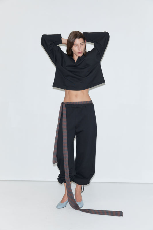 A person stands against a white background, wearing a loose black long-sleeve top, the Side Pleat Pant in black—a wide-leg cotton style with a long plaid belt reminiscent of Deiji Studios' relaxed designs—and light blue shoes, with arms raised and hands touching their head.