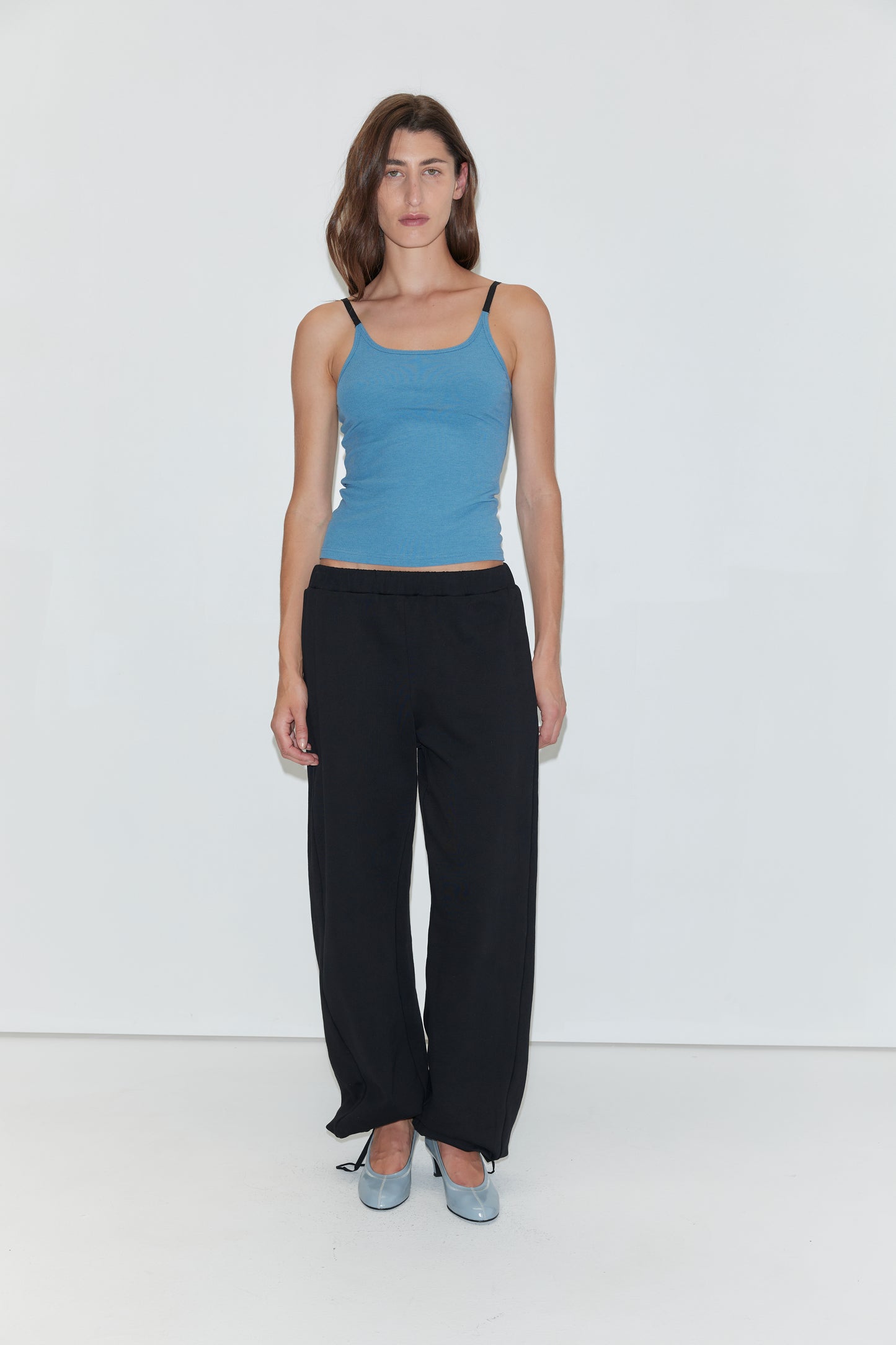 A woman with long brown hair wears a blue tank top, paired with the Deiji Studios Side Pleat Pant in black and light blue shoes. She stands against a plain white background, looking directly at the camera with a neutral expression.