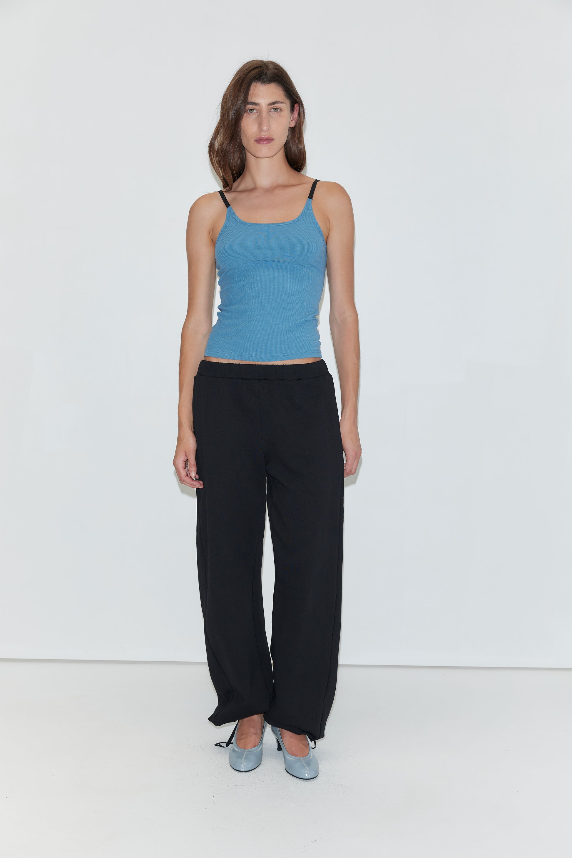 A woman with long brown hair wears a blue tank top, paired with the Deiji Studios Side Pleat Pant in black and light blue shoes. She stands against a plain white background, looking directly at the camera with a neutral expression.