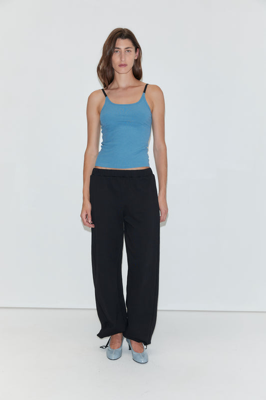 A woman with long brown hair wears a blue tank top, paired with the Deiji Studios Side Pleat Pant in black and light blue shoes. She stands against a plain white background, looking directly at the camera with a neutral expression.