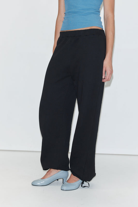 A person wearing a blue sleeveless top, Deiji Studios Side Pleat Pant - Black with an elastic drawcord, and light blue heels stands against a white background. Only the person’s torso and legs are visible.