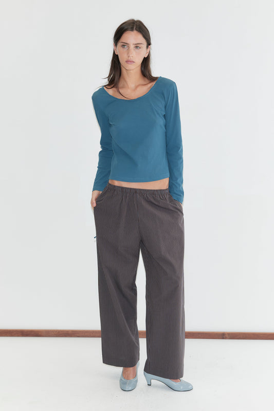 A woman with long dark hair stands against a plain white background wearing the Ease Trouser - Brown Blue Stripe by Deiji Studios, dark brown wide-leg trousers with a fine blue pinstripe, with a teal long-sleeve top and pale blue heels.