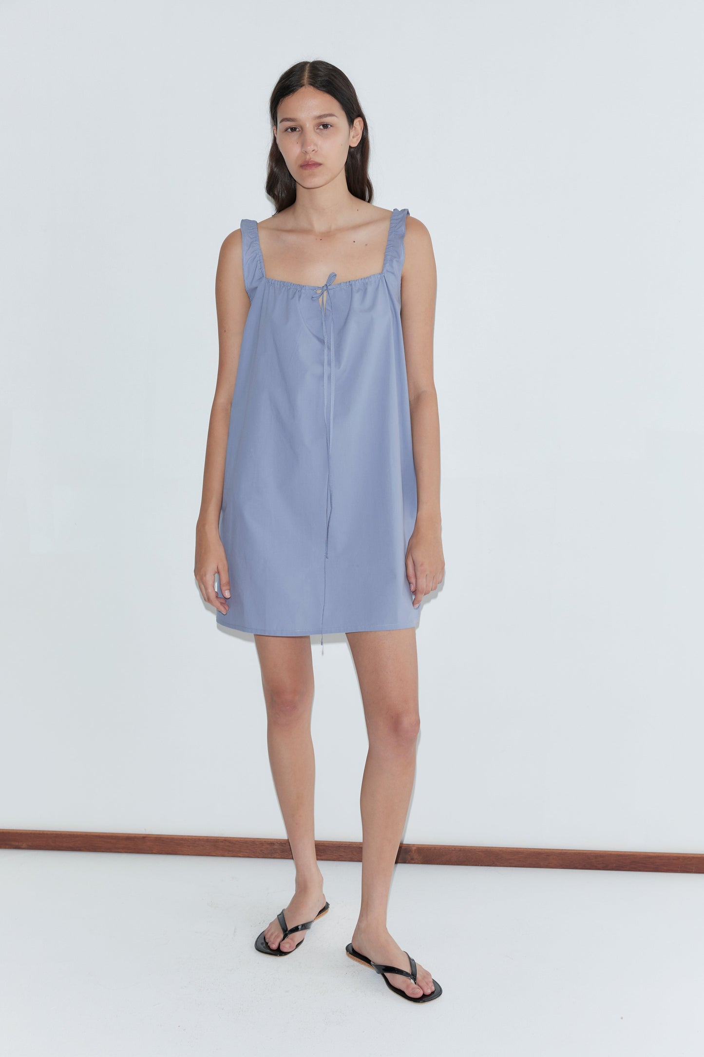 A woman with long dark hair and a neutral expression wears the mini-length Bow Dress - Iris by Deiji Studios, featuring ruffled straps, paired with black flip-flops against a plain white background.