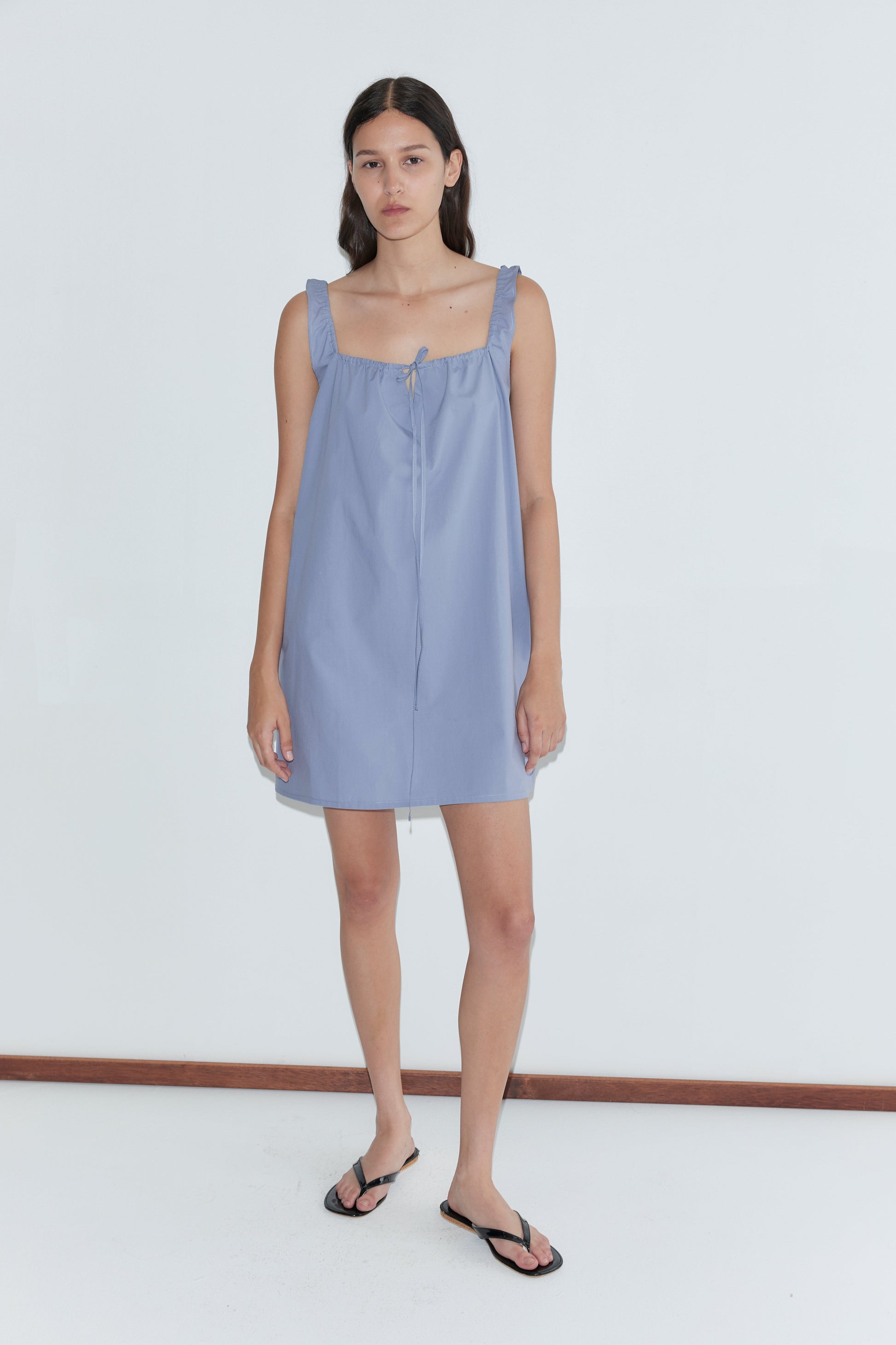 A woman with long dark hair and a neutral expression wears the mini-length Bow Dress - Iris by Deiji Studios, featuring ruffled straps, paired with black flip-flops against a plain white background.