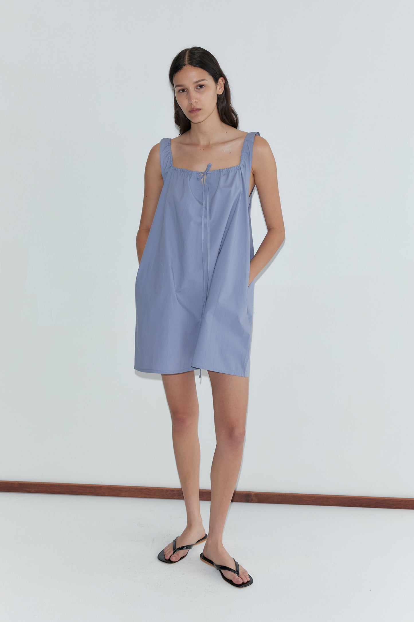 A woman with long, straight dark hair and a neutral expression stands against a white wall, wearing the Bow Dress - Iris by Deiji Studios, a loose mini-length dress with pockets, paired with black flip-flops.