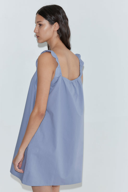 A woman with long dark hair wears the Deiji Studios Bow Dress - Iris, a loose-fitting, sleeveless mini dress with ruffled straps. She stands against a plain light background, facing away and turning her head to the side.