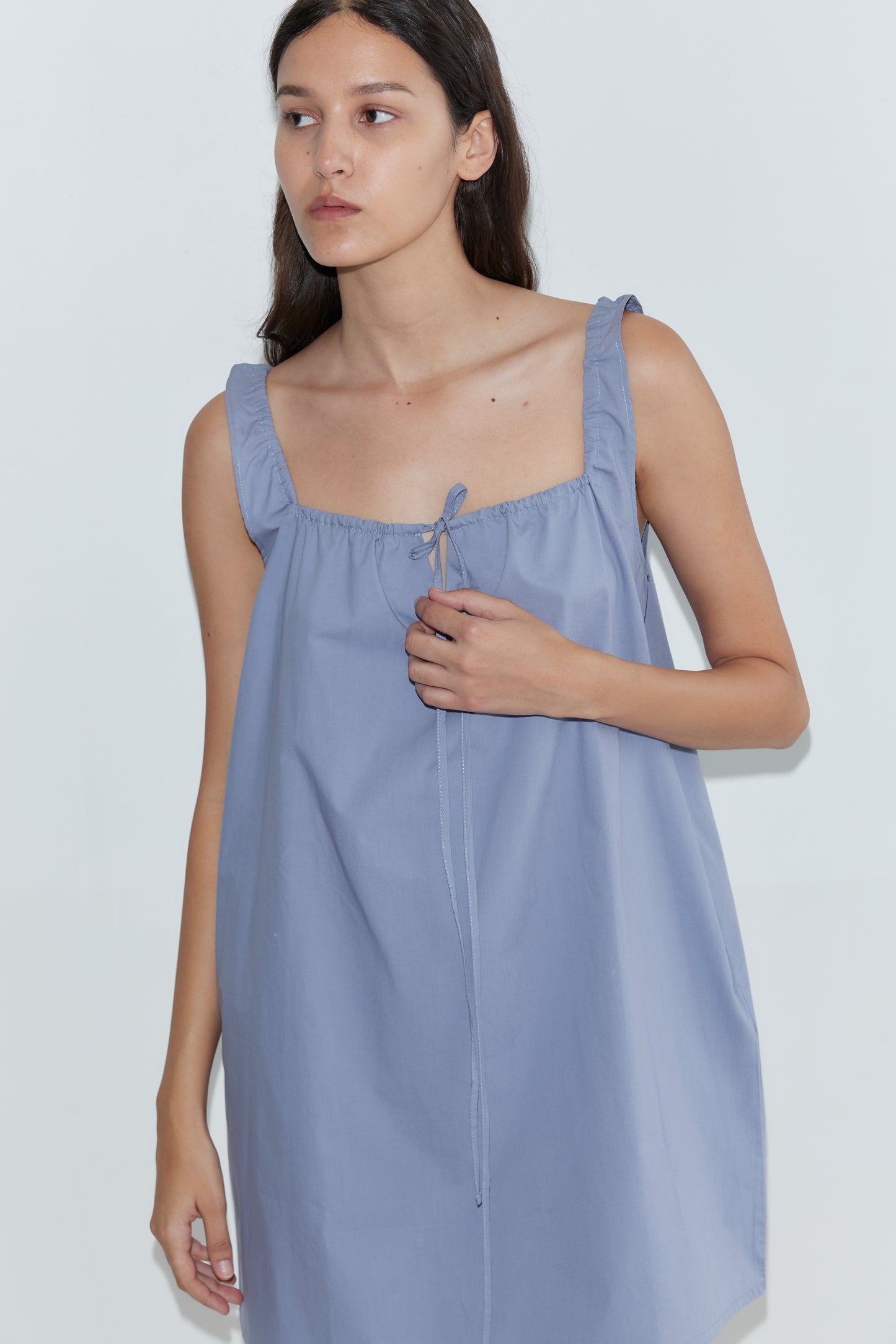 A woman with long dark hair wears the Bow Dress - Iris by Deiji Studios, a loose mini dress with ruffled straps and a tie neckline, standing against a plain white background.
