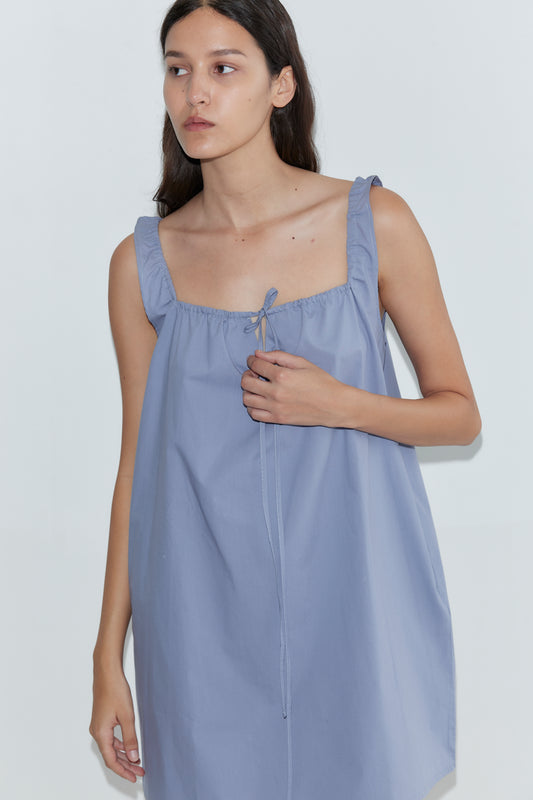 A woman with long dark hair wears the Bow Dress - Iris by Deiji Studios, a loose mini dress with ruffled straps and a tie neckline, standing against a plain white background.
