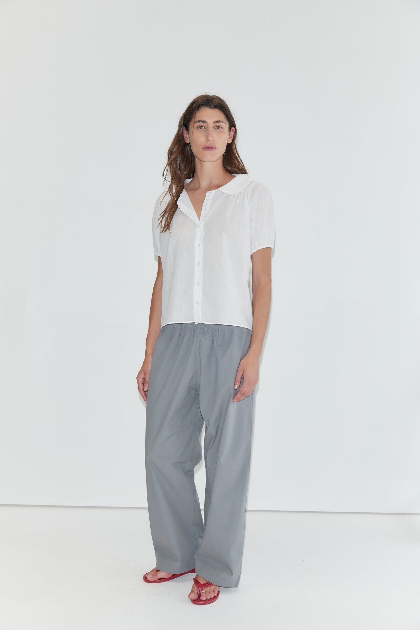 A person stands against a white background wearing the Petal Top - White, a short-sleeve cotton voile top with a delicate peter pan collar, paired with loose gray pants and red sandals.