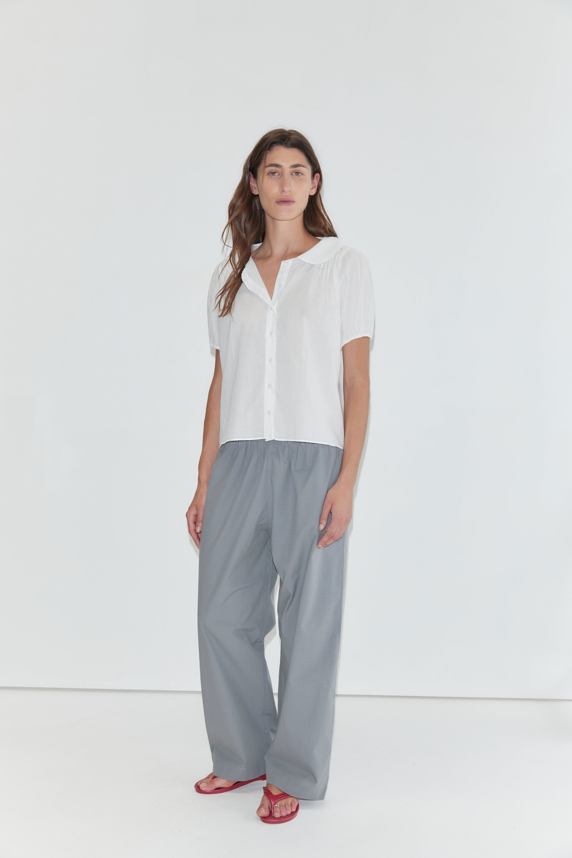 A person stands against a white background wearing the Petal Top - White, a short-sleeve cotton voile top with a delicate peter pan collar, paired with loose gray pants and red sandals.