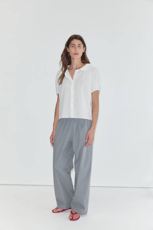 A person stands against a white background wearing the Petal Top - White, a short-sleeve cotton voile top with a delicate peter pan collar, paired with loose gray pants and red sandals.