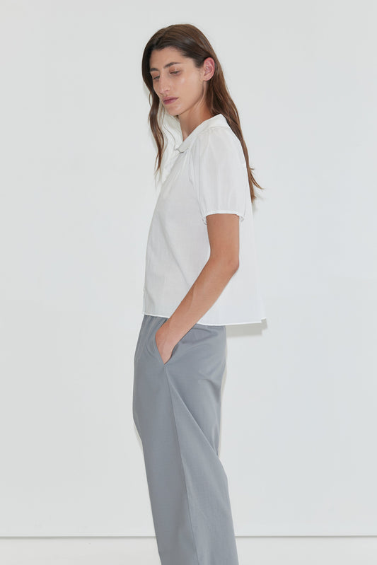 Person with long dark hair stands in profile against a plain white background, eyes closed and hands in pockets, wearing the Petal Top - White from Deiji Studios—a short-sleeve cotton voile blouse featuring a subtle peter pan collar—paired with light gray pants.