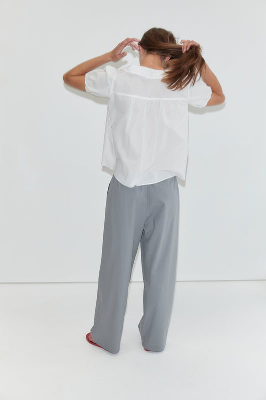 A person stands facing a white wall, holding their brown hair in a ponytail. They wear the Petal Top in White, featuring a subtle Peter Pan collar, paired with loose gray pants and red shoes. The photo is taken indoors with a minimal background, echoing the understated elegance of Deiji Studios’ style.