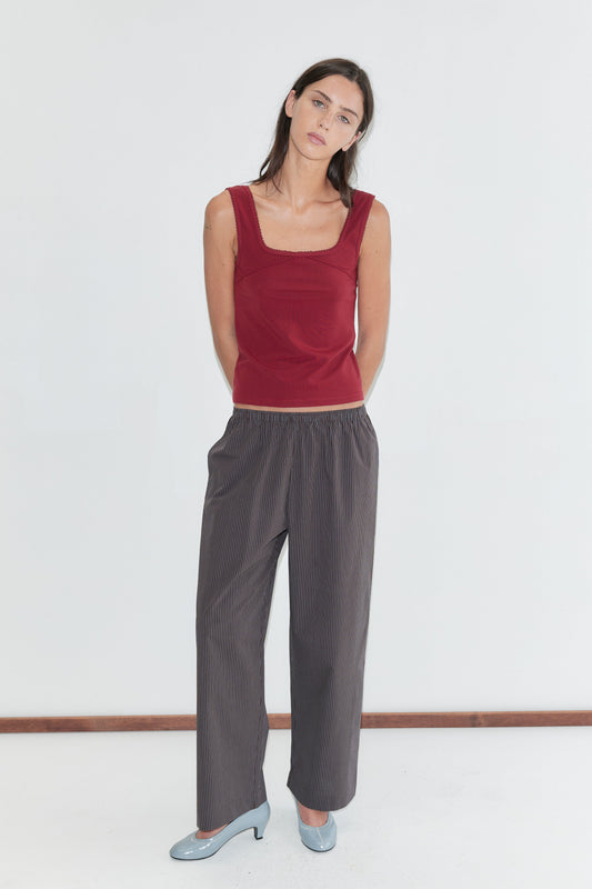 A woman with long dark hair stands against a plain white background wearing the Ease Trouser - Brown Blue Stripe by Deiji Studios, dark brown wide-leg trousers with a fine blue pinstripe, with a red square-neck tank and pale blue heels.