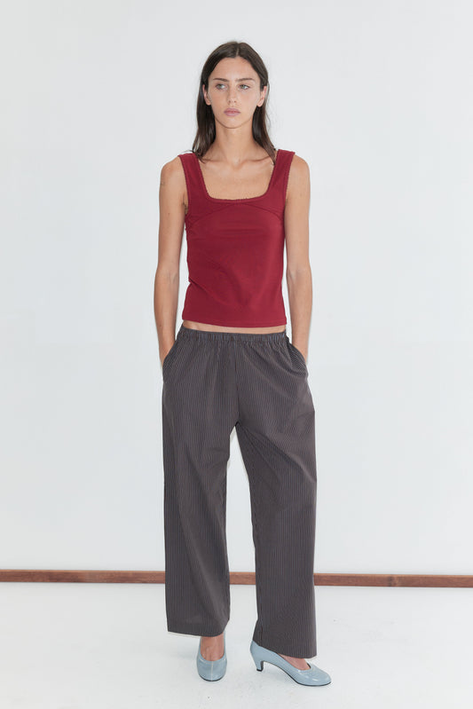 A woman with long dark hair stands against a plain white background wearing the Ease Trouser - Brown Blue Stripe by Deiji Studios, dark brown wide-leg trousers with a fine blue pinstripe, paired with a red square-neck tank and pale blue heels.