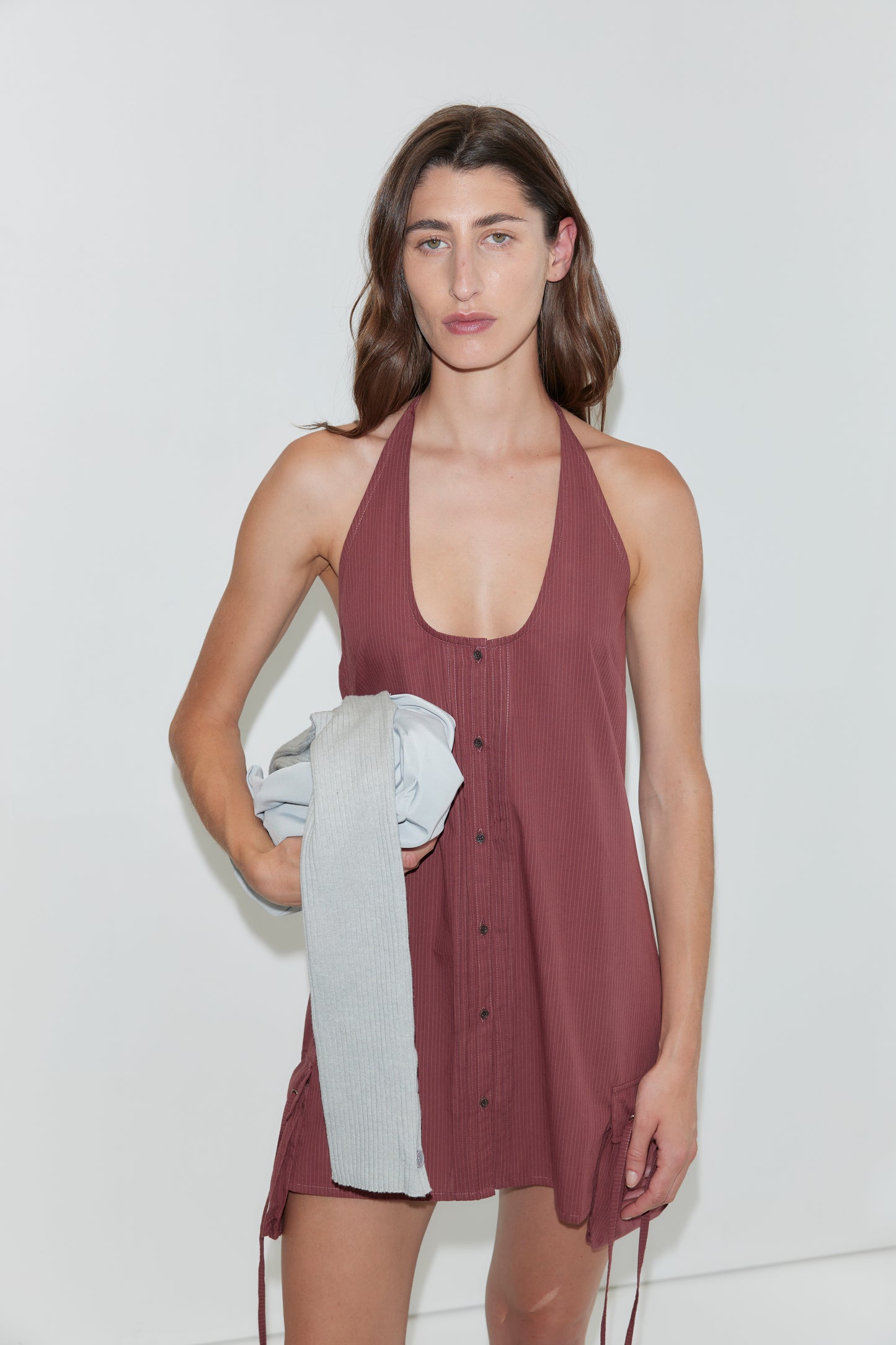 A person with long brown hair, wearing a sleeveless, button-up Pocket Halter Dress in Rust Stripe, stands against a white background holding a folded gray garment in one hand.