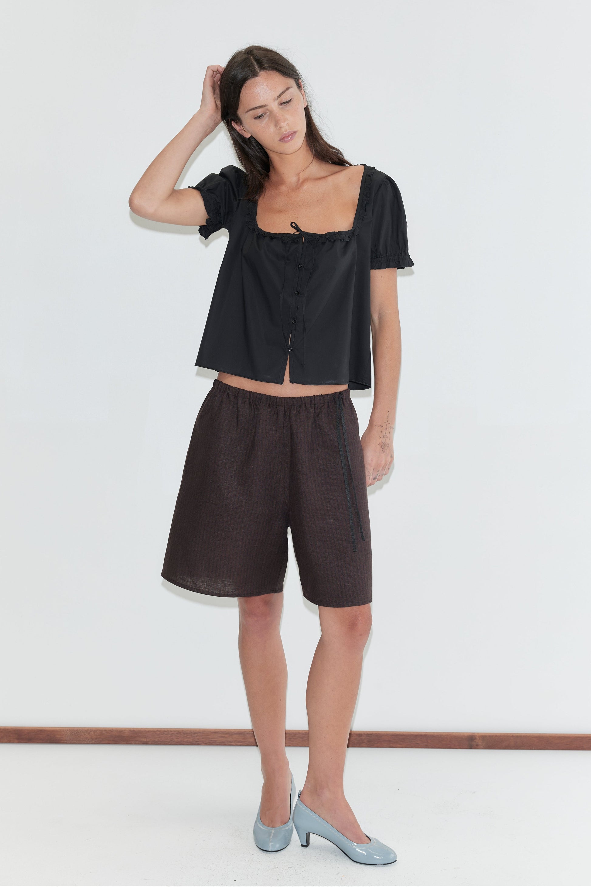 A woman with long dark hair stands against a plain white background wearing the Half Short - Brown Black Stripe by Deiji Studios, knee-length dark brown striped linen shorts, with a black flounced top and pale blue heels.