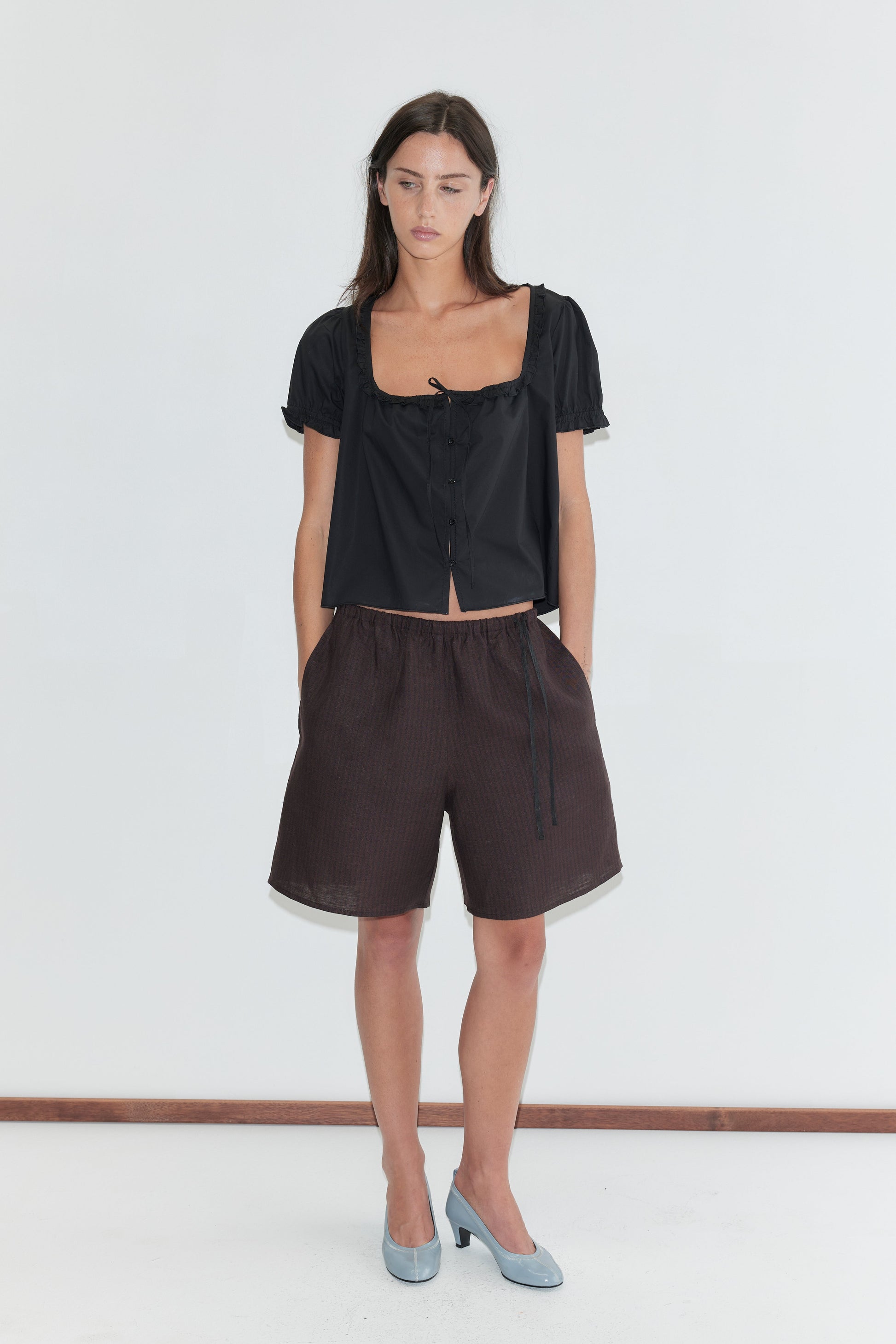 A woman with long dark hair stands against a plain white background wearing the Flounced Top - Black by Deiji Studios, a black top with a frilled square neckline, front tie and puff sleeves, with brown striped linen shorts and pale blue heels.