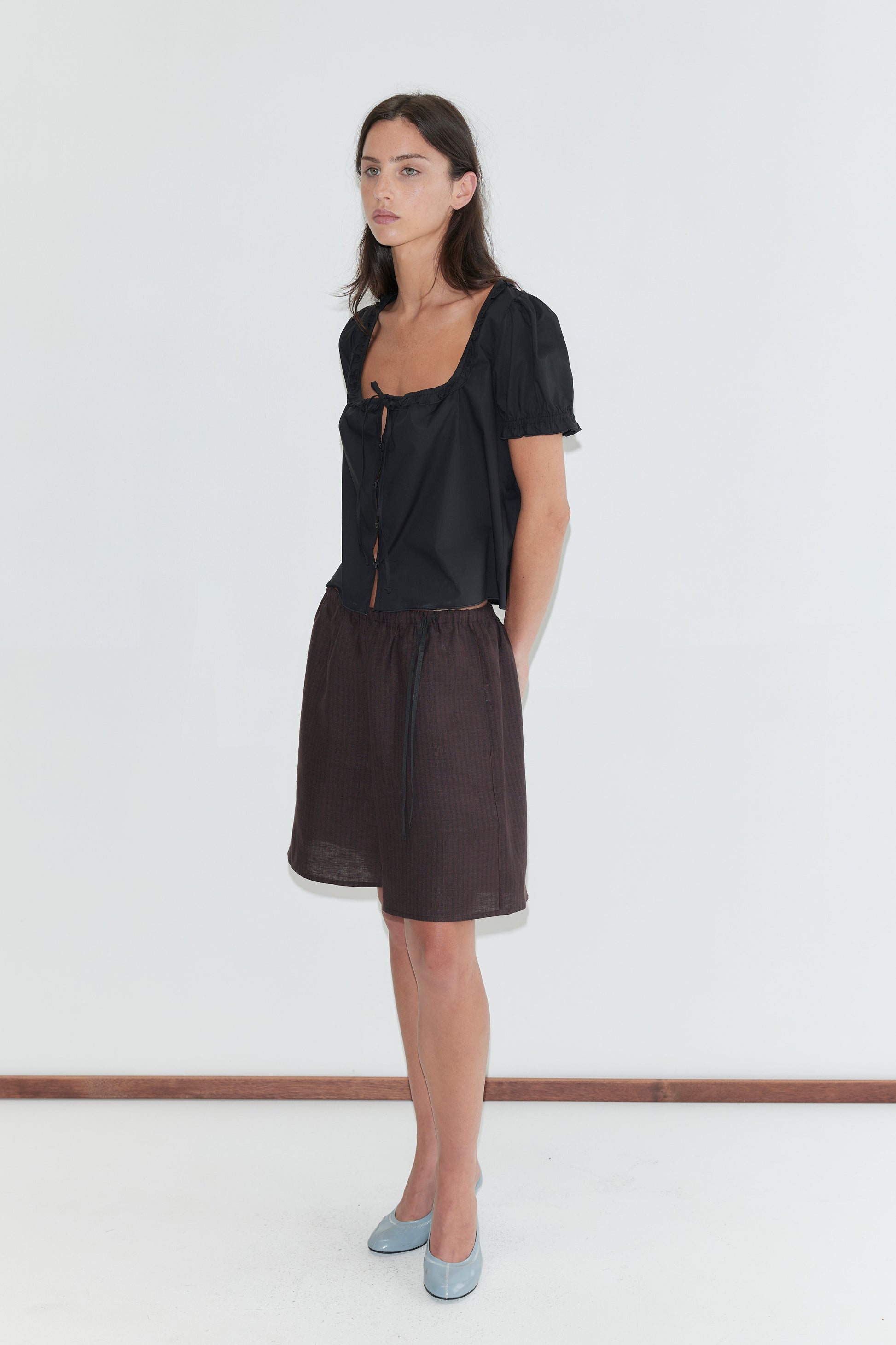 A woman with long dark hair stands against a plain white background wearing the Flounced Top - Black by Deiji Studios, a black top with a frilled square neckline and puff sleeves, with brown striped linen shorts and pale blue heels.