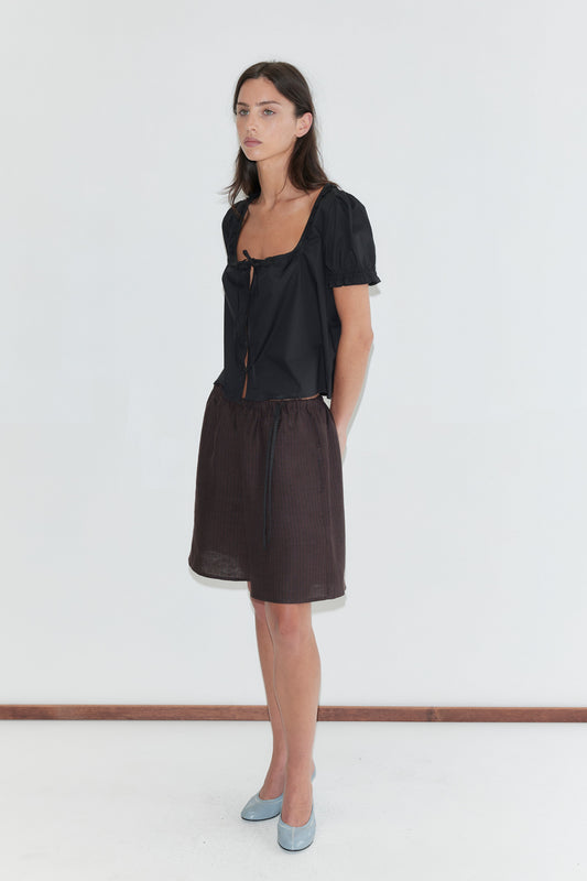 A woman with long dark hair stands against a plain white background wearing the Flounced Top - Black by Deiji Studios, a black top with a frilled square neckline and puff sleeves, with brown striped linen shorts and pale blue heels.