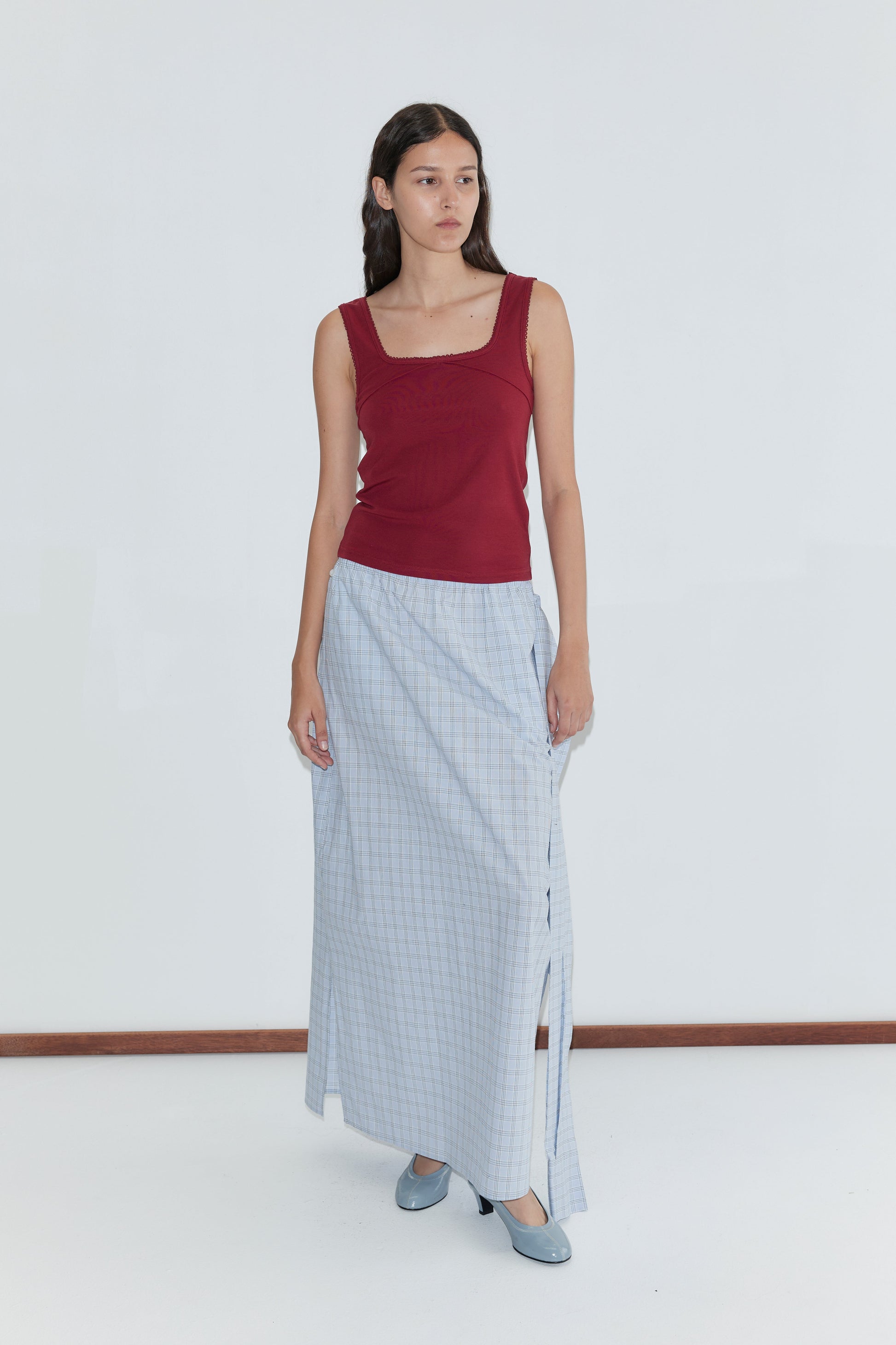 A woman with long dark hair stands calmly against a white wall, wearing Deiji Studios’ Piping Tank in Crimson, a long light blue checked skirt with side slit, and matching shoes.