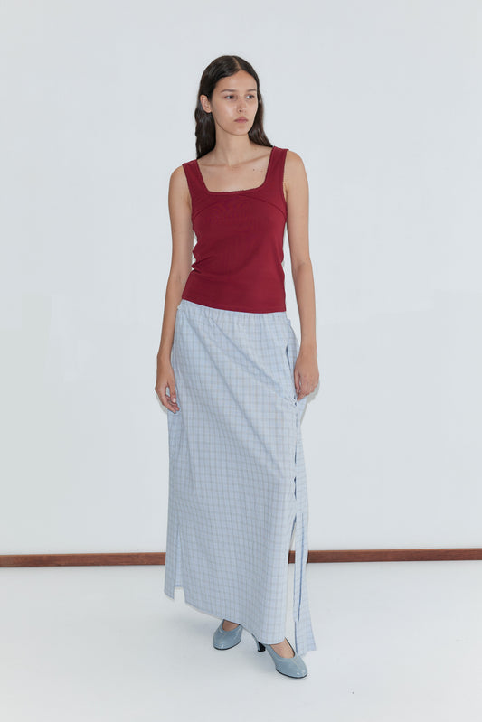 A woman with long dark hair stands calmly against a white wall, wearing Deiji Studios’ Piping Tank in Crimson, a long light blue checked skirt with side slit, and matching shoes.