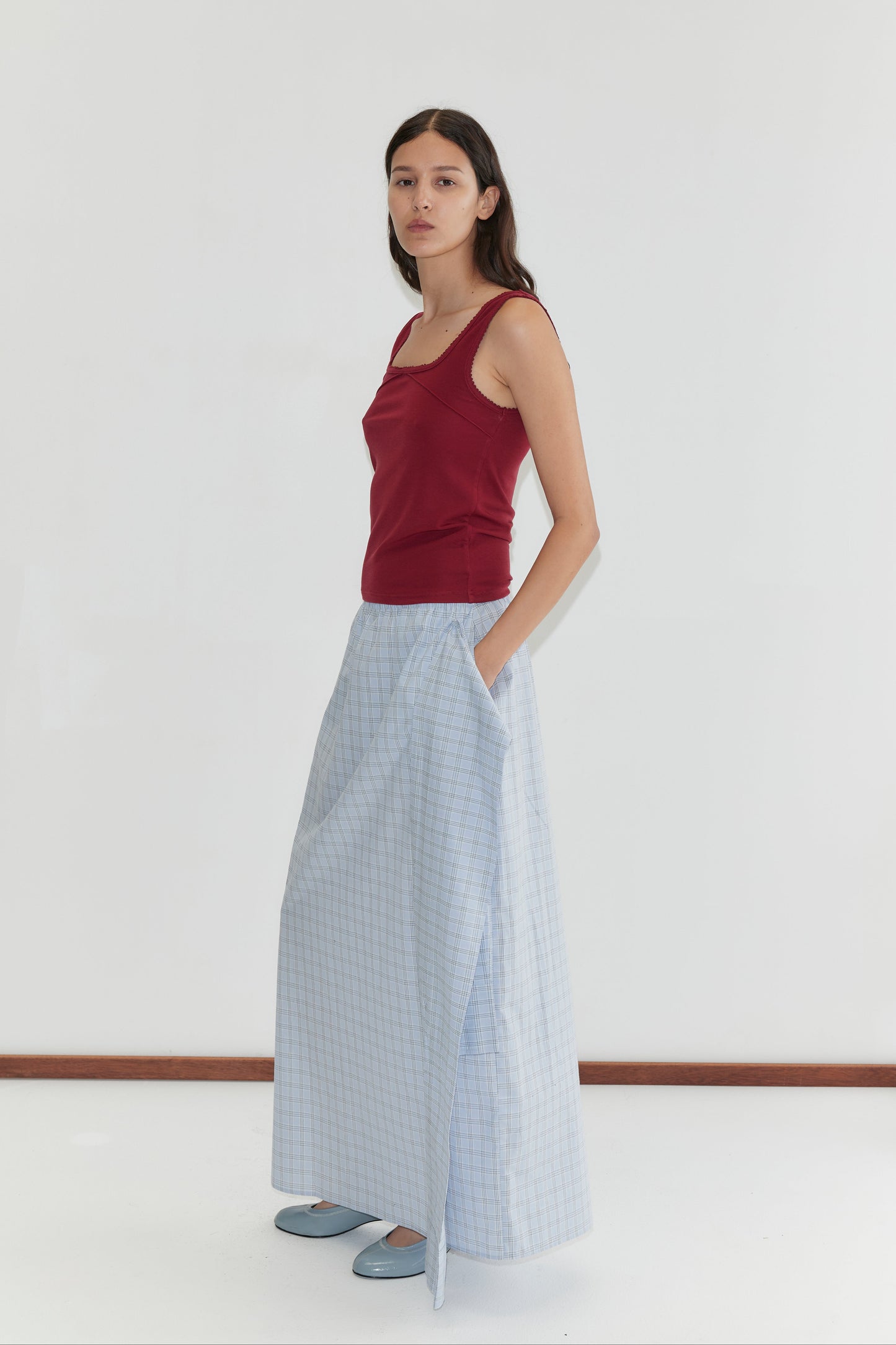 A woman with long brown hair and a neutral expression stands against a white background, wearing the Piping Tank - Crimson with a square neckline from Deiji Studios, paired with a light blue checked skirt and matching shoes.