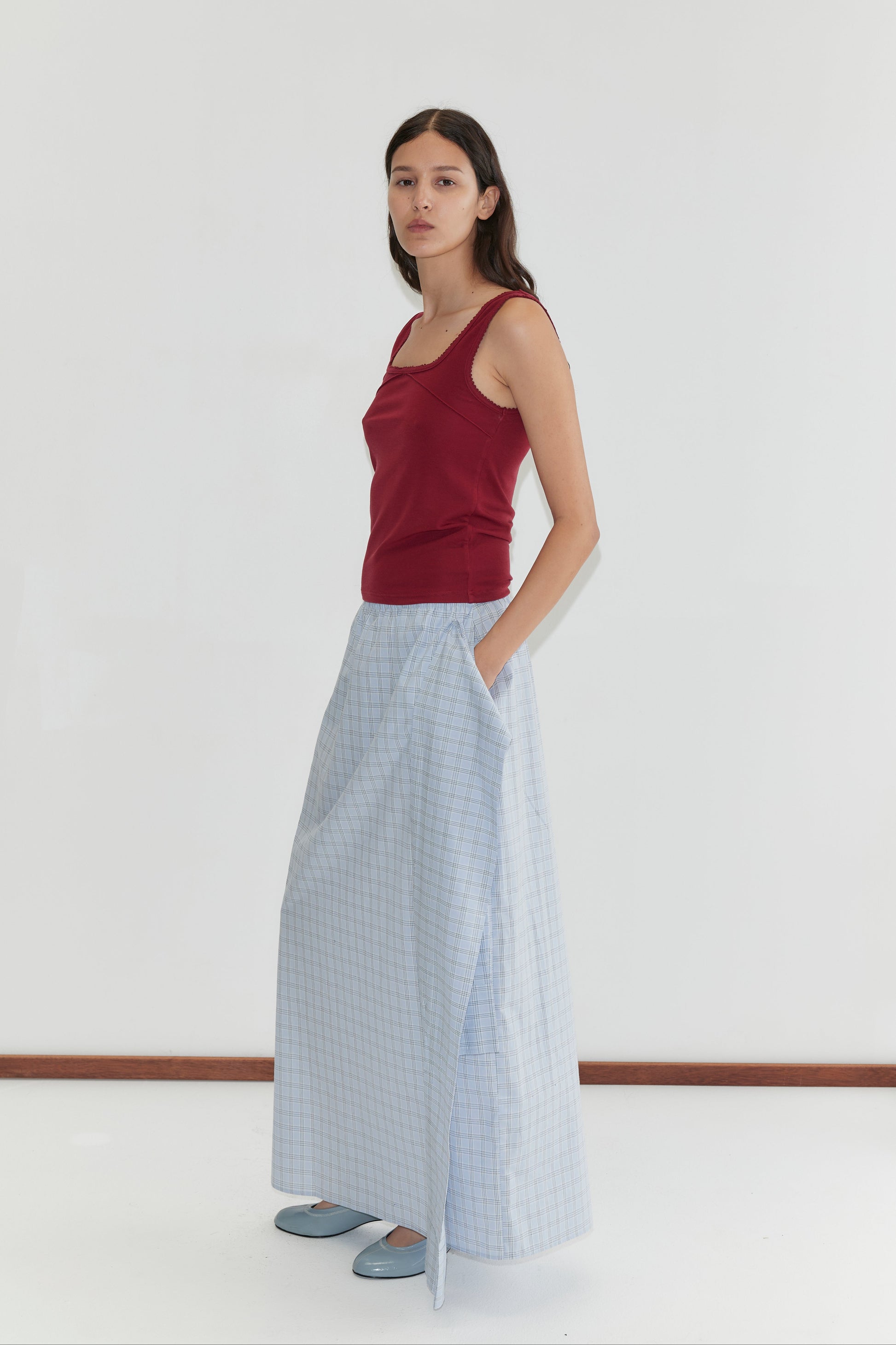 A woman with long brown hair and a neutral expression stands against a white background, wearing the Piping Tank - Crimson with a square neckline from Deiji Studios, paired with a light blue checked skirt and matching shoes.